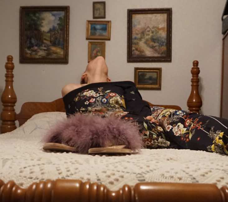 a woman in a blue pant set with purple feather shoes on a bed with a lace bed spread 