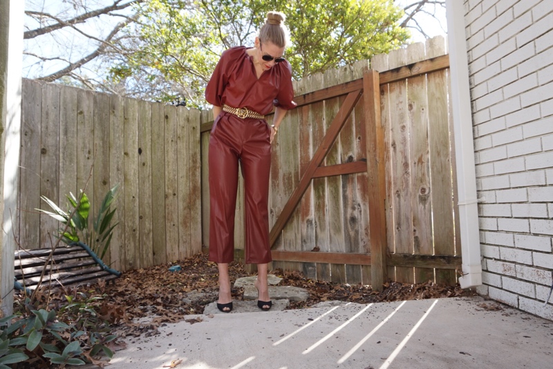 a woman in a brown faux leather top and pants with a gold belt and black peep toe pumps by a fence and a white brick wall