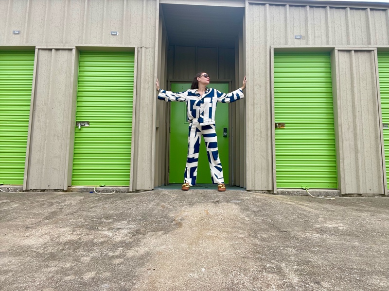 a woman in a blue and white linen pant and white long sleeve pant set in front of green doors and concrete walls  