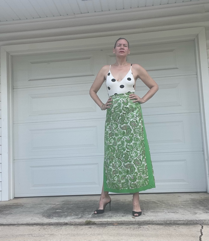a woman in a white with black  polka dot s body suit  and a green with embroidered panels maxi skirt and black heeled mules 