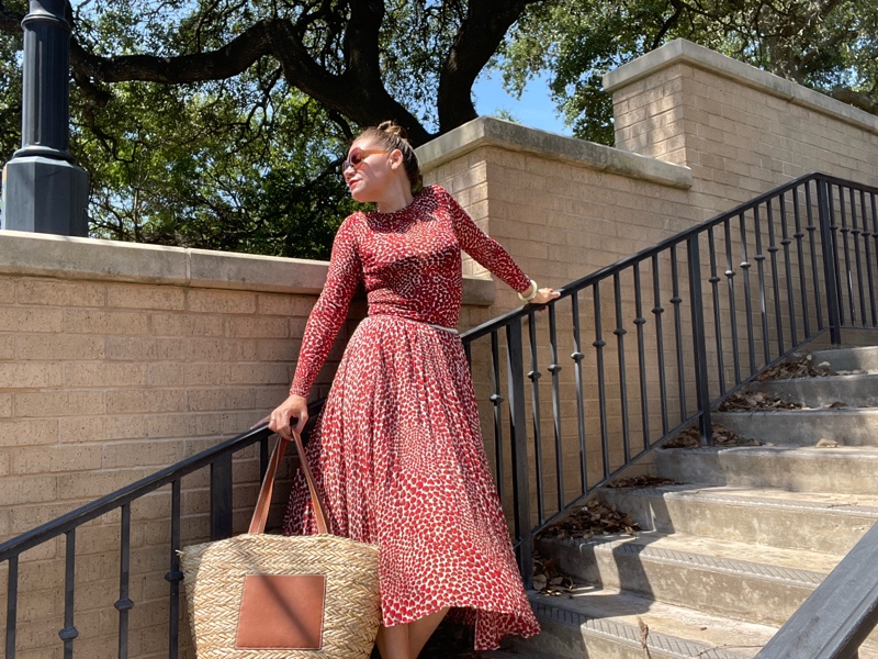 a woman in  strawberry print top and skirt holding a straw and leather tote with gold mule