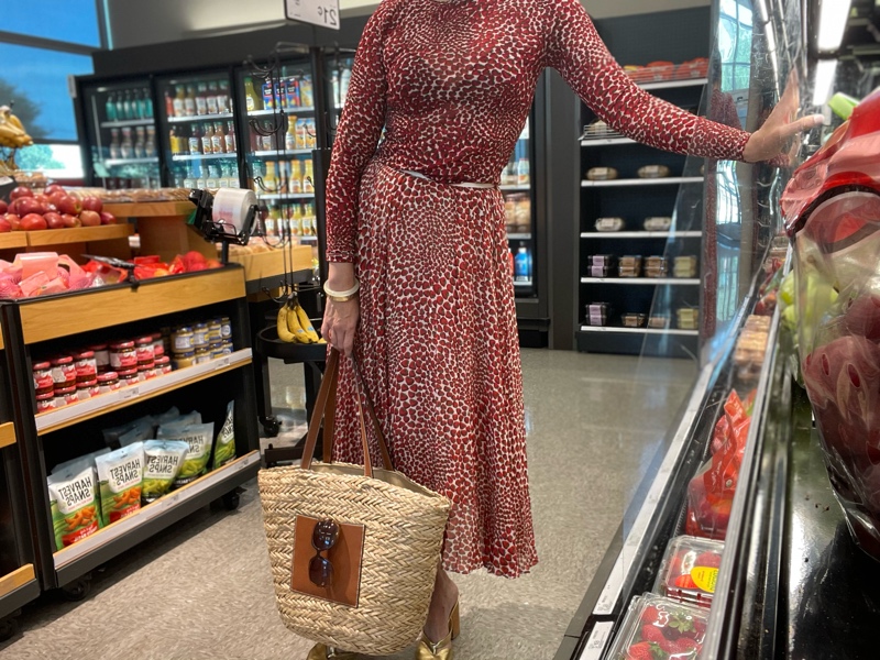 a woman in a strawberry topi and skirt set with a straw and leather tote and gold mules in a grocery store