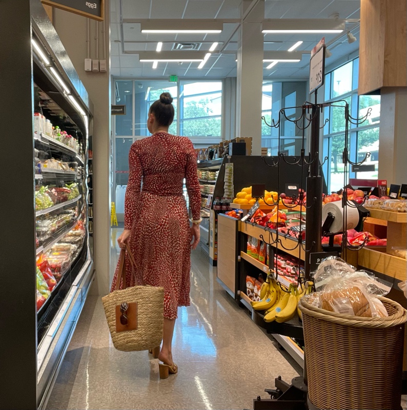 a woman in a strawberry topi and skirt set with a straw and leather tote and gold mules in a grocery store